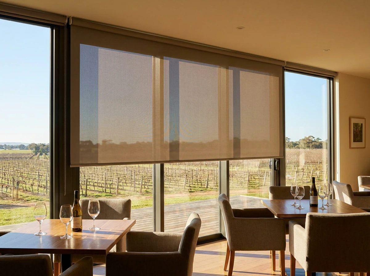 Flat sunscreen roller blind lowered over a tasting-room glass wall with vineyard rows visible through the mesh